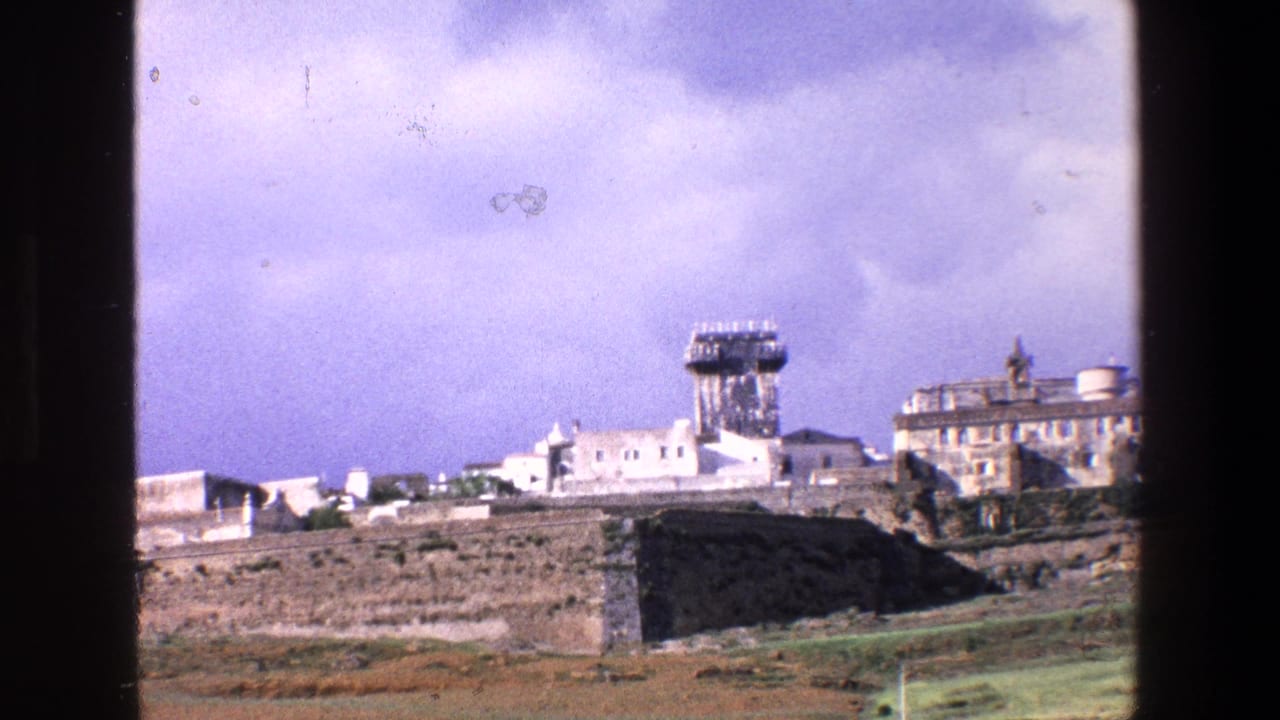 1967 Palermo home-movie frame showing a distant stone fortification, white buildings and a prominent tower under a pale purplish sky, partially framed by a dark window edge.