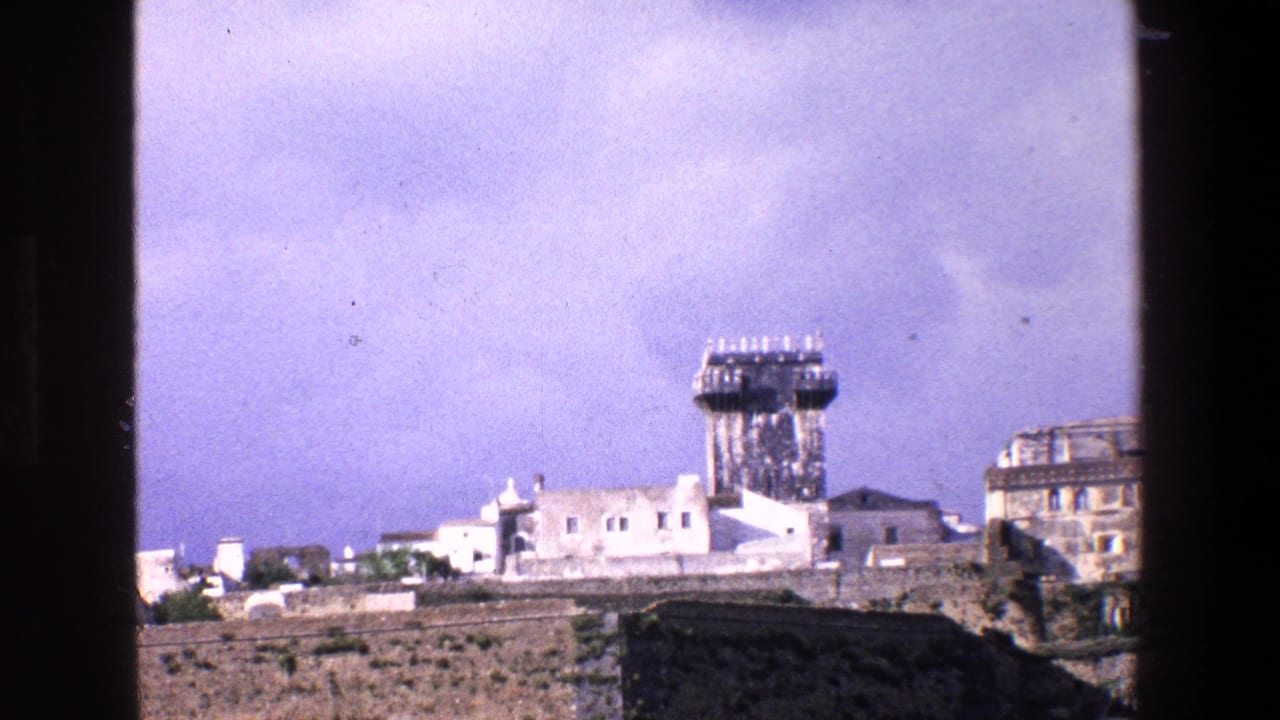 1967 Palermo travel footage frame with a distant fortress wall, low white buildings and a prominent tower, with the scene divided by the dark vertical window frame.