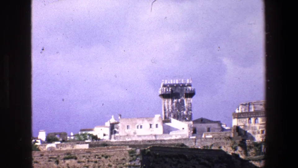1967 Palermo 8mm frame showing a closer visitor-shot view of stone walls, white buildings and a prominent tower, with soft focus and visible film grain.
