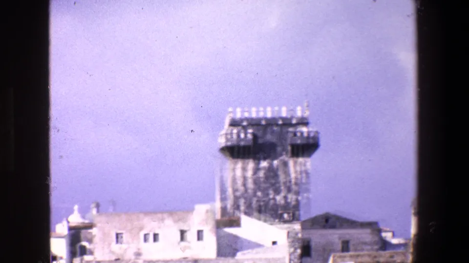 Zoomed 1967 Palermo home-movie frame centering on the prominent tower and adjacent white buildings, with the dark edge of the window frame still visible at the side.