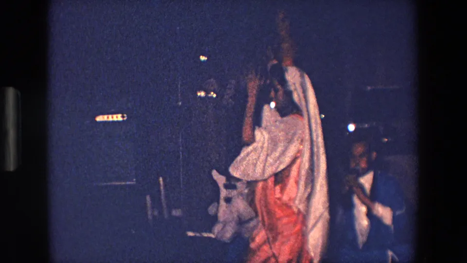 1967 Palermo frame of a robed female performer or participant moving through a dim nighttime festival scene, with people, lights, and dark street surroundings behind her.