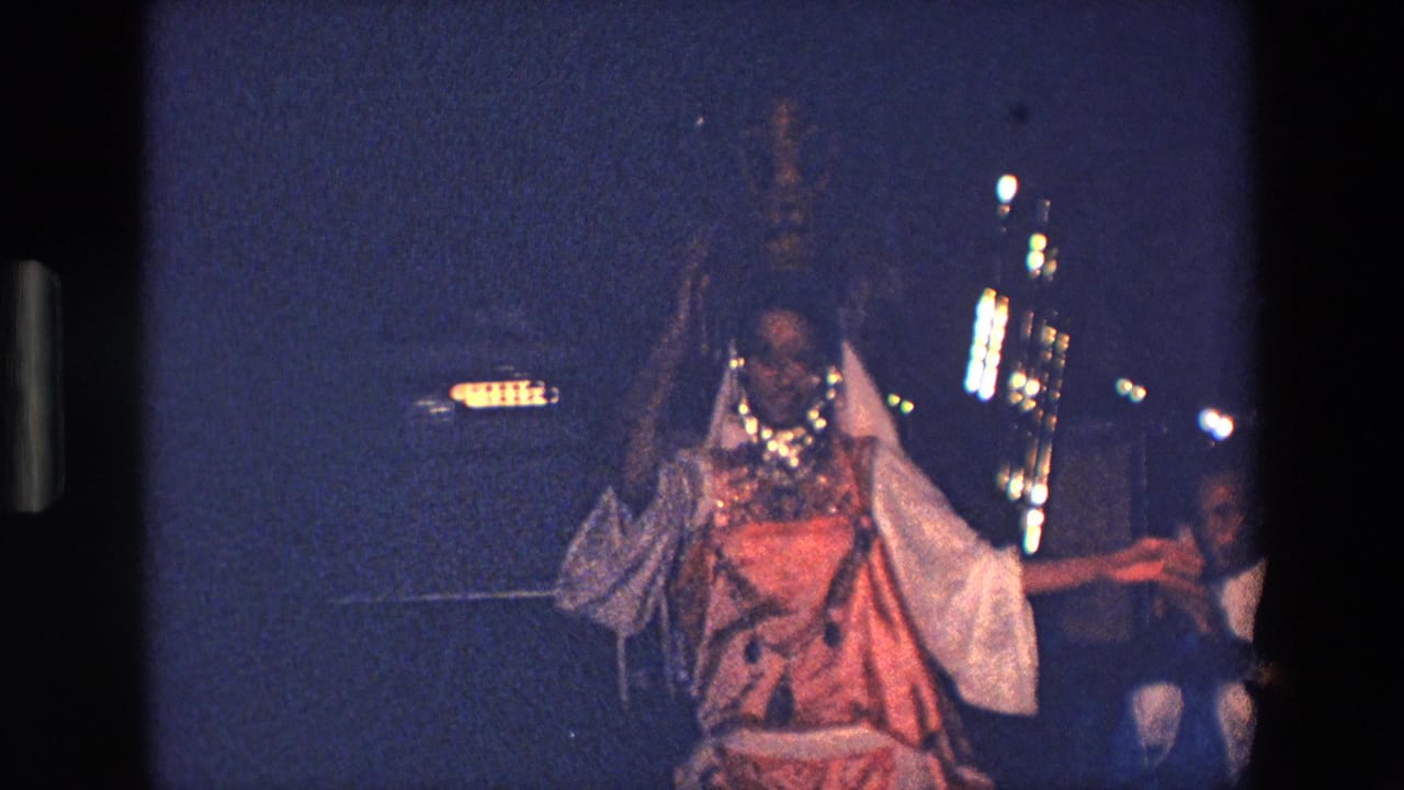 In Palermo, Italy, 1967 home-movie footage, a woman in a light veil and orange-white costume balances a tall ornate vessel on her head during a nighttime performance.