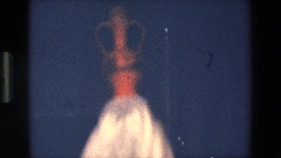 A blurred low-light frame shows the woman moving through the nighttime performance, with the tall vessel still visible above her head and her pale costume catching the exposure.