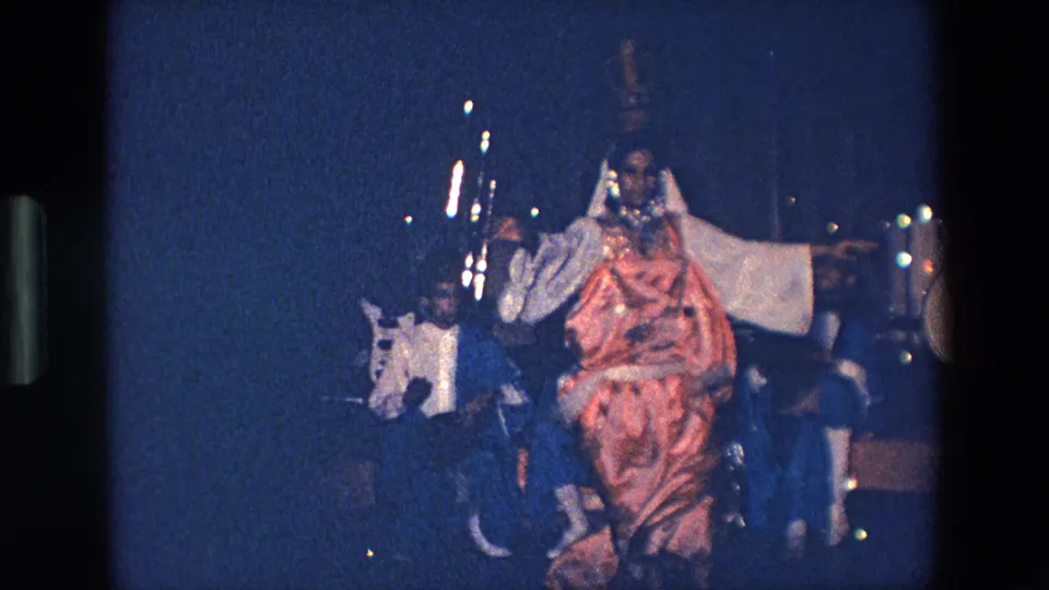 A wide 8mm home-movie view of the Palermo performance shows the dancer in red costume extending her arm while the band remains visible in the background.