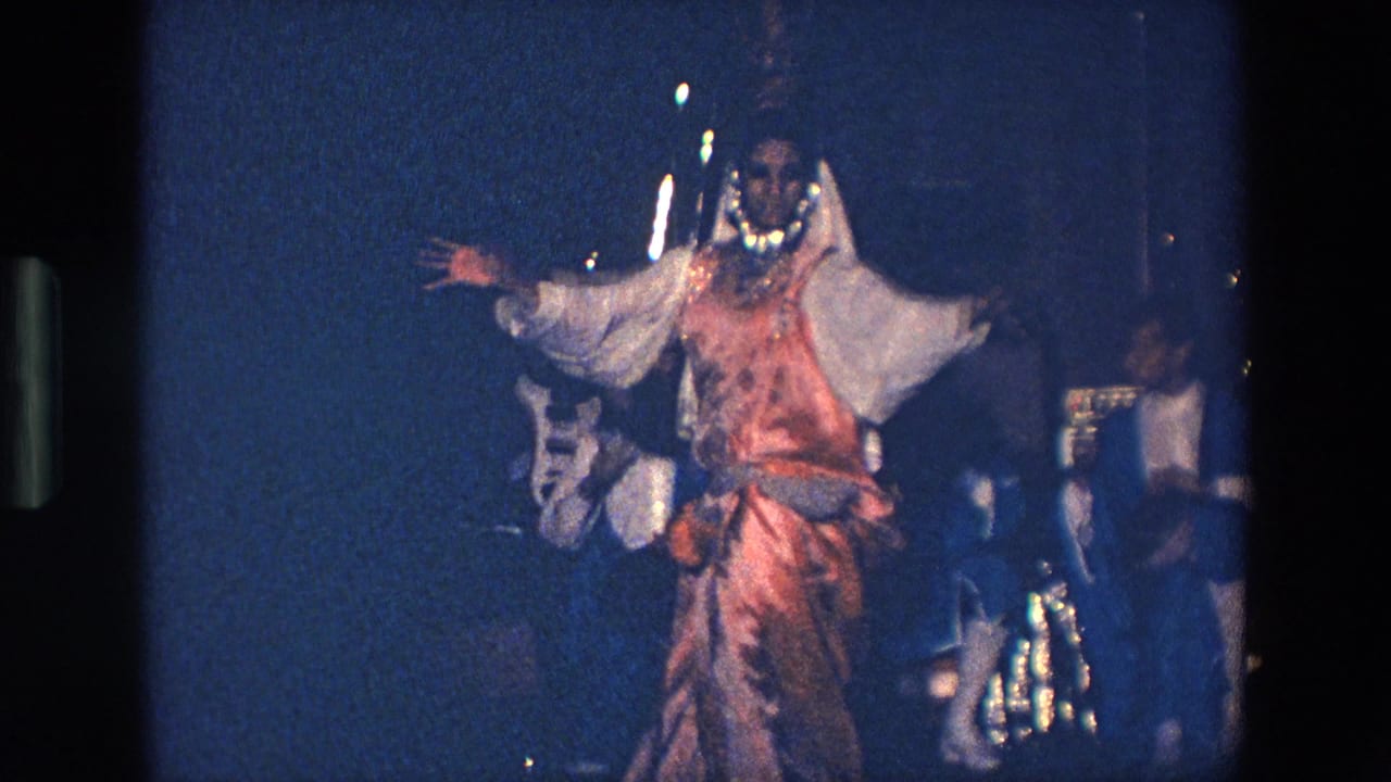 In Palermo, Italy in 1967, a woman in an orange and white Indian-inspired costume stands with arms raised during a dimly lit stage dance.