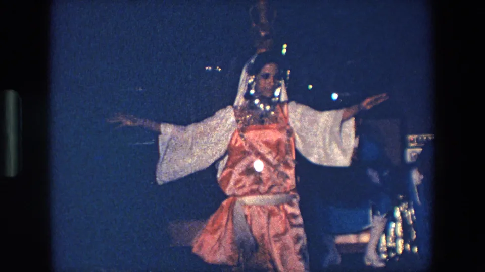 A woman in a bright orange and white dance costume holds a side-facing arm pose in the Palermo 1967 stage performance footage.