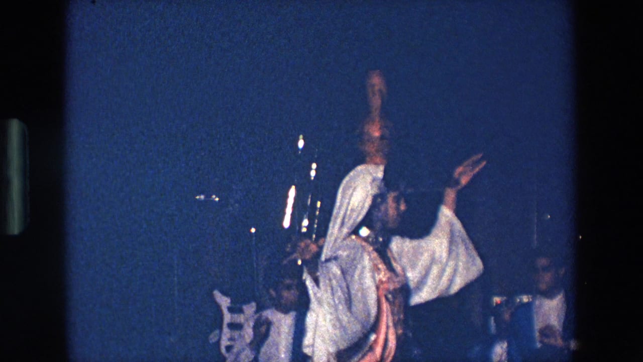 In metadata-supported Palermo, Italy, 1967, a woman in white and brown costume dances under blue stage lighting with a clay amphora balanced on her head while musicians appear behind her.