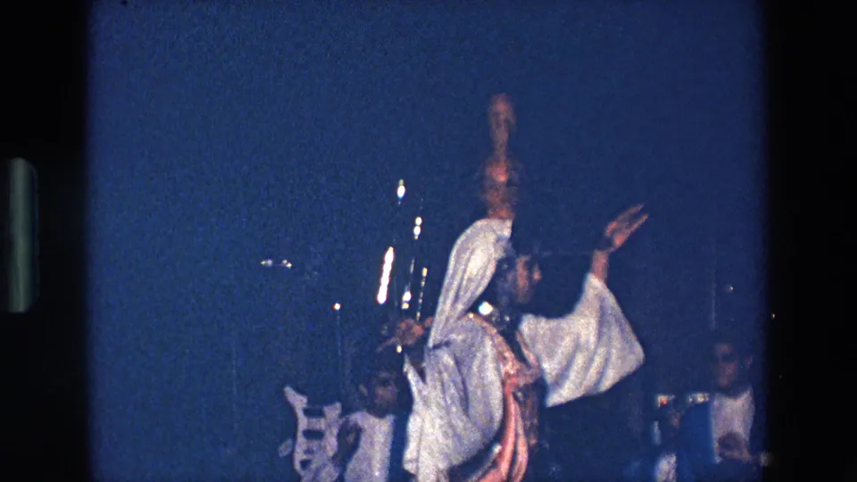 In metadata-supported Palermo, Italy, 1967, a woman in white and brown costume dances under blue stage lighting with a clay amphora balanced on her head while musicians appear behind her.