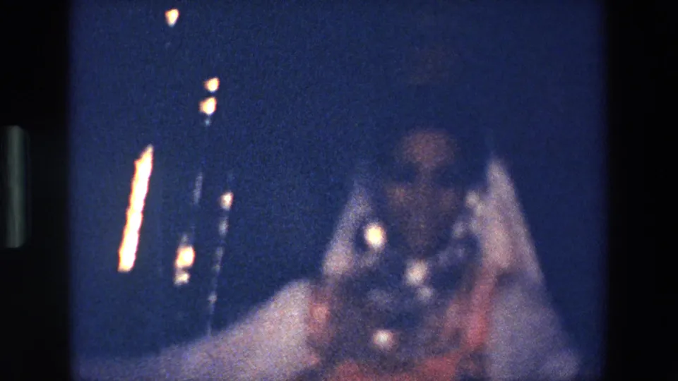 The home-movie camera catches a heavily blurred low-light moment of the dancer’s movement, with bright reflections and the white costume visible against the dark stage.