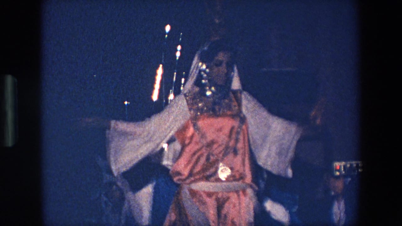 In supported 1967 Palermo footage, a woman in a pink costume with wide white sleeves dances with arms raised while a band is partly visible behind her.