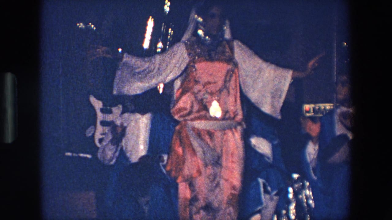 Costumed woman dances with arms extended as a guitarist stands behind her on a dark stage.