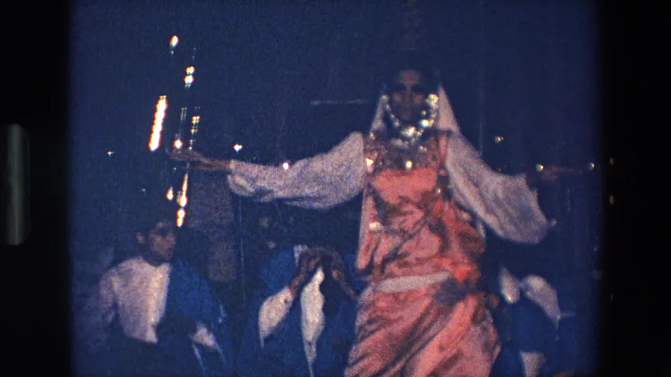 The 1967 Palermo home-movie frame shows the dancer facing forward in a pink costume and white veil, with musicians and instruments partially visible behind her.
