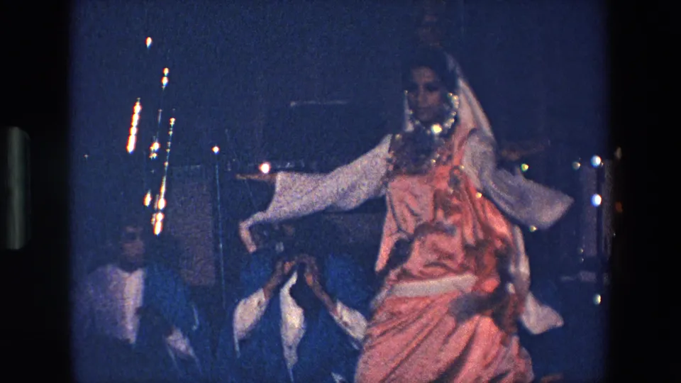 A soft 8mm frame from the Palermo performance shows the dancer mid-motion, her pink costume and veil blurred by movement as musicians sit behind her.