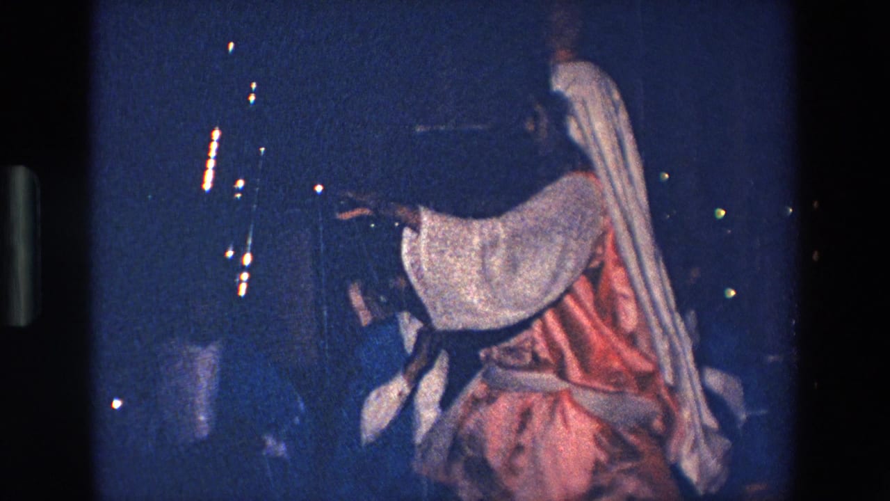In 1967 Palermo home-movie footage, a robed performer with a white head covering and red garment gestures in dim blue night light near a small crowd.