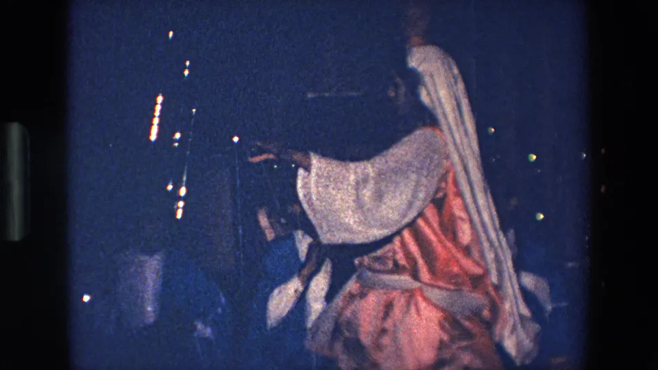 In 1967 Palermo home-movie footage, a robed performer with a white head covering and red garment gestures in dim blue night light near a small crowd.