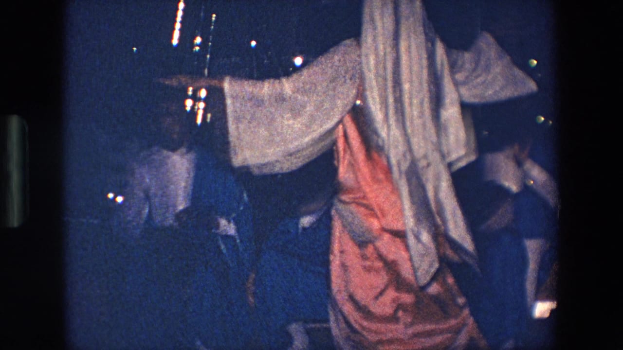 A traditionally dressed performer, seen from behind in 1967 Palermo footage, extends an arm toward spectators under dim outdoor lighting.