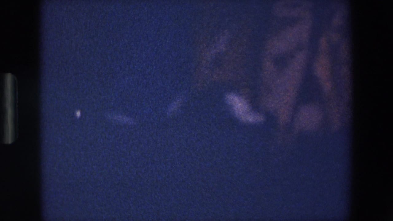 1967 Palermo home-movie frame with pink skirt fabric and part of a woman’s lower leg seen through heavy grain and blue-purple low light.