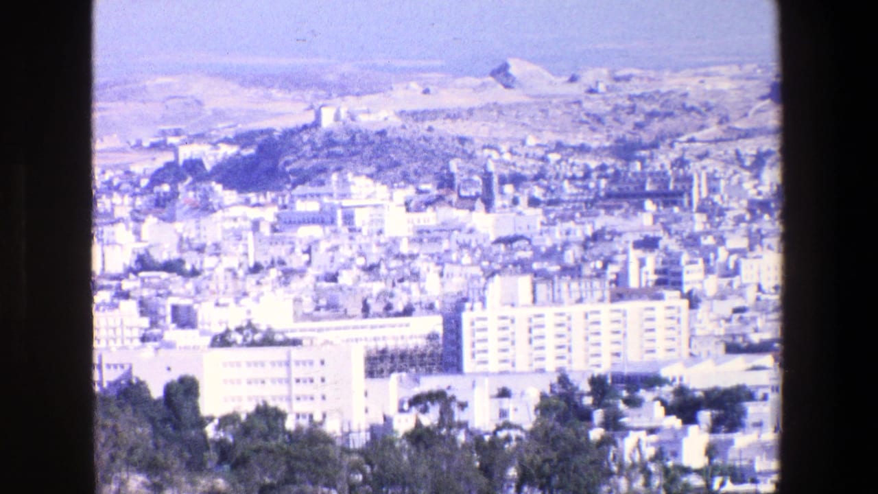 1967 8mm home-movie frame from Palermo showing a bright elevated city panorama with pale buildings, apartment blocks, hills, and a distant hilltop structure.