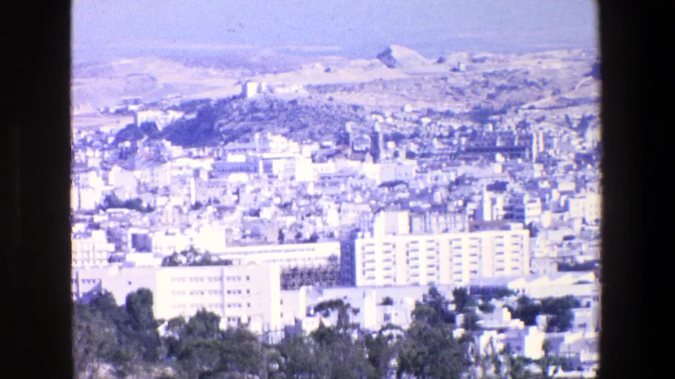 1967 8mm home-movie frame from Palermo showing a bright elevated city panorama with pale buildings, apartment blocks, hills, and a distant hilltop structure.