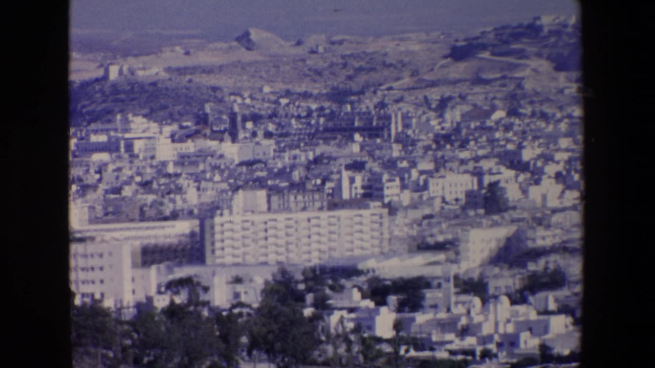 Panoramic cityscape with apartment blocks, pale buildings, and hills in the background.