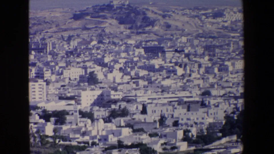 1967 Palermo 8mm frame showing the city and hillside from an elevated viewpoint, with pale buildings, possible roads or terraces, and a distant hilltop structure.