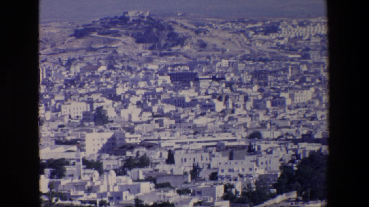1967 8mm home-movie frame showing Palermo from above, with tightly packed pale buildings, a distant hilltop landmark, and soft blue film color.
