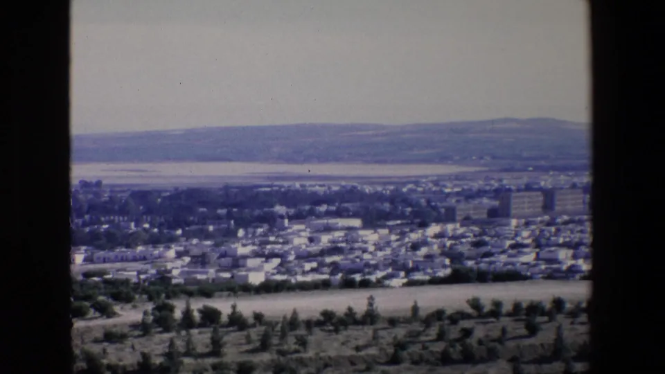 1967 8mm frame from Palermo showing foreground trees and fields, low city buildings, a band of coastal water, and mountains in the distance.