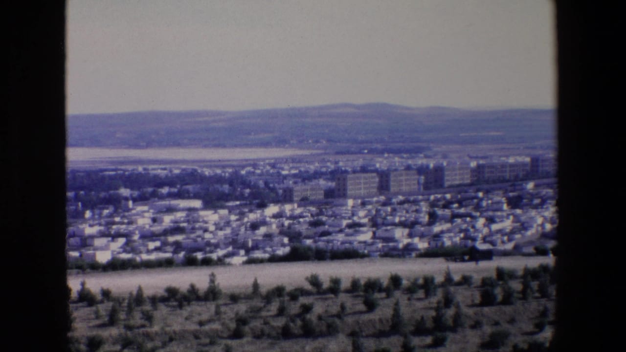 1967 Palermo home-movie frame showing a hazy panoramic cityscape with dense low buildings, taller apartment blocks, distant hills, and dark structural edges in the foreground.