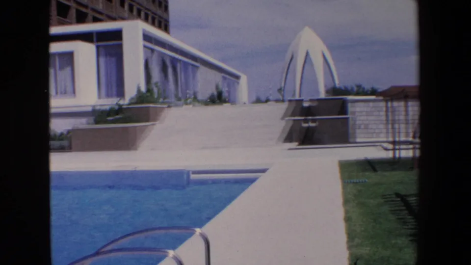 1967 Palermo home-movie frame showing a modern poolside exterior with a bright blue swimming pool, white deck, adjacent house, lawn, and a distinctive white arched structure.