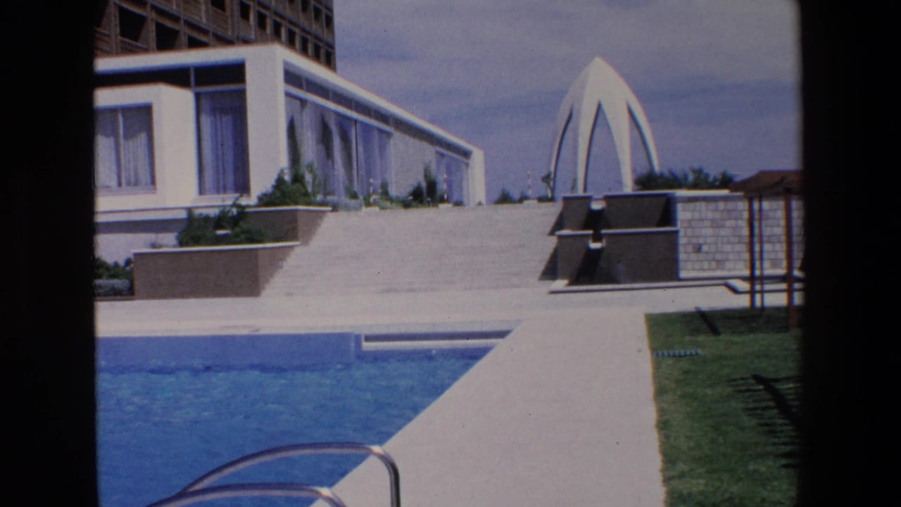 1967 Palermo travel footage frame showing a blue hotel pool, concrete walkway, landscaped terrace, and white modernist building with a sculptural feature in the background.