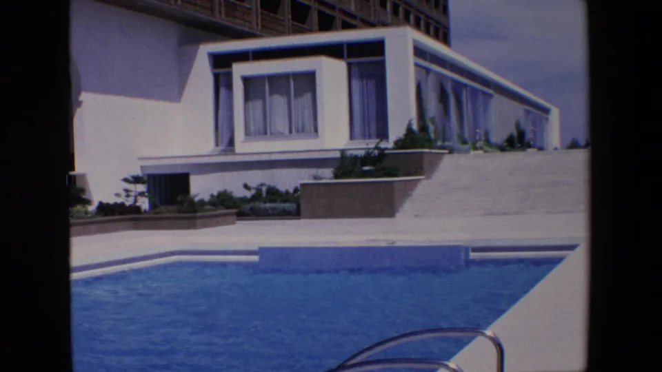 1967 Palermo frame showing the swimming pool edge, a white modernist hotel block, planted terrace beds, and a broad sloped walkway rising toward the building.