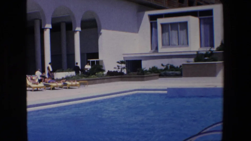 1967 Palermo resort frame with guests on lounge chairs near arched colonnades, a blue swimming pool in the foreground, and the modernist hotel facade behind them.