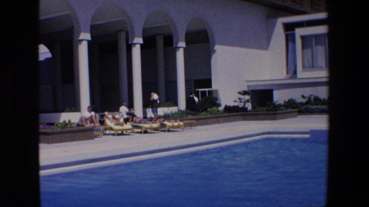 1967 Palermo home-movie frame showing guests on yellow lounge chairs beside a swimming pool under a shaded colonnade with white pillars.