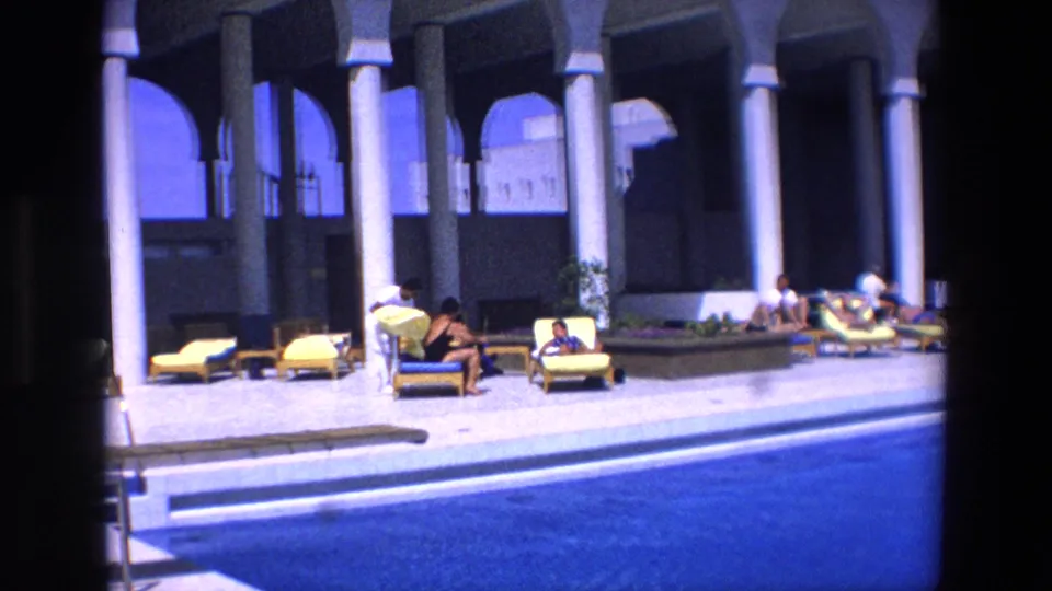 1967 8mm frame of sunbathers and seated guests beside a resort swimming pool, with arched architecture and tall columns in view.