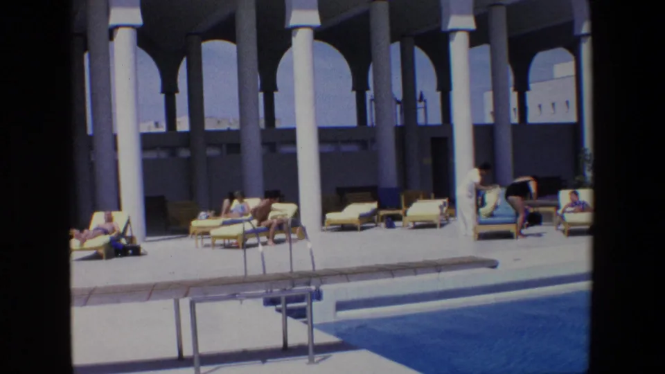 Wide 1967 Palermo resort pool view with guests scattered among lounge chairs, a ladder at the pool edge, and a shaded colonnade behind them.