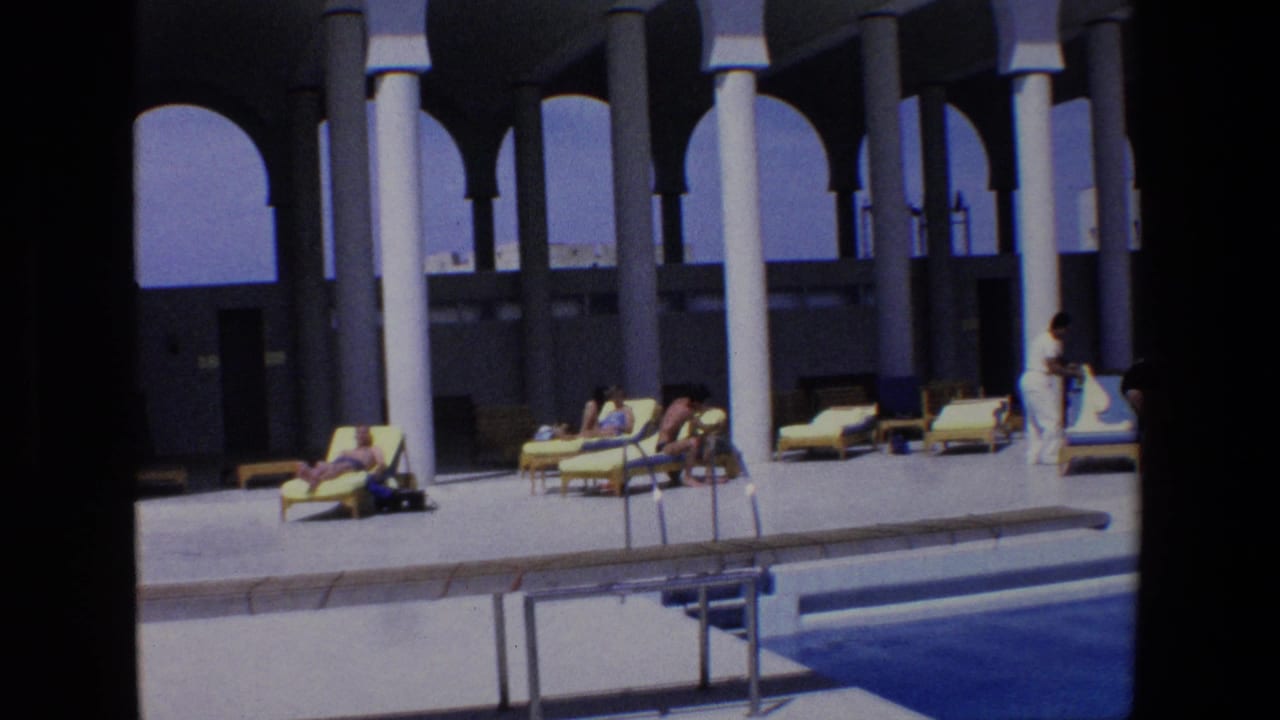 1967 Palermo home-movie frame showing several people in swimwear resting on lounge chairs by a pool beneath a white-columned portico.