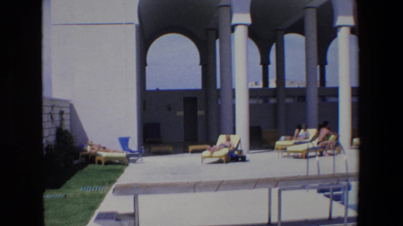 People sit and recline on deck chairs near a pool and arched portico.
