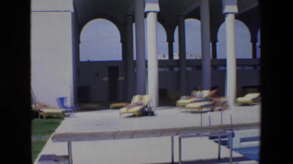1967 Palermo footage frame showing a sunlit poolside area with lounge chairs, towels, seated figures, white portico columns, and arched openings.