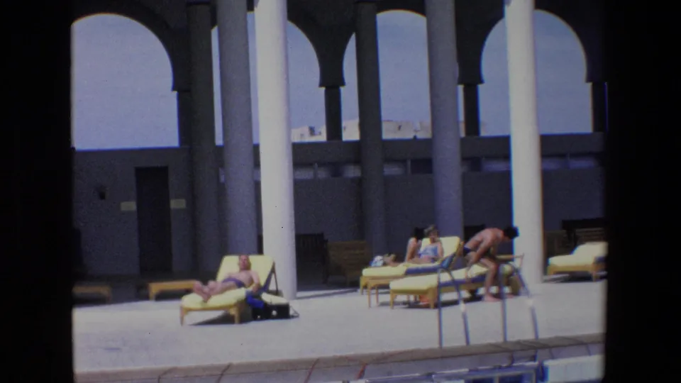 1967 Palermo home-movie frame with people in swimwear beside a pool, including a man bending near lounge chairs under a shaded white-columned portico.