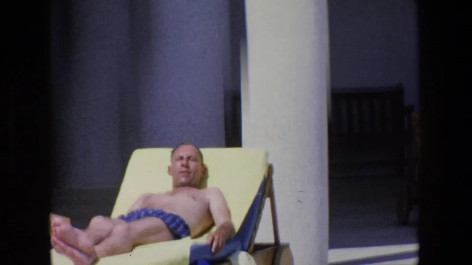 Medium close 1967 Palermo home-movie frame of a man resting on a yellow lounge chair, framed by a white column and darker patio background.