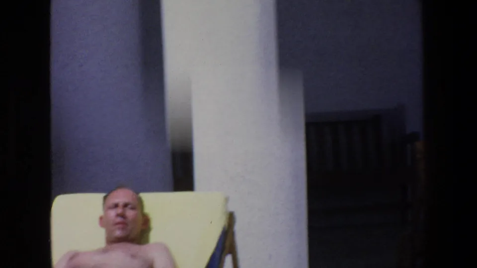 Tight 1967 Palermo frame showing the reclining man's face and upper body on a yellow lounge chair, with a white column and shaded bench in the background.