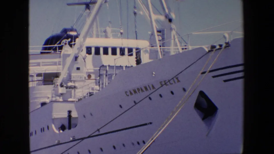 1967 Palermo home-movie frame looking up at the Campania Felix hull and bow, with readable ship lettering, portholes, rigging, deck structures, and pier railings.