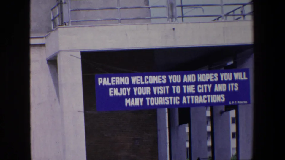 1967 Palermo port frame showing a blue welcome banner beneath a concrete structure, with white text greeting visitors to the city and its tourist attractions.