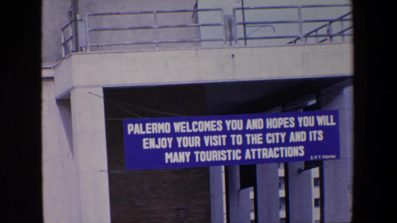 1967 Palermo travel footage frame showing a blue welcome banner with white lettering beneath a concrete overpass and railings.