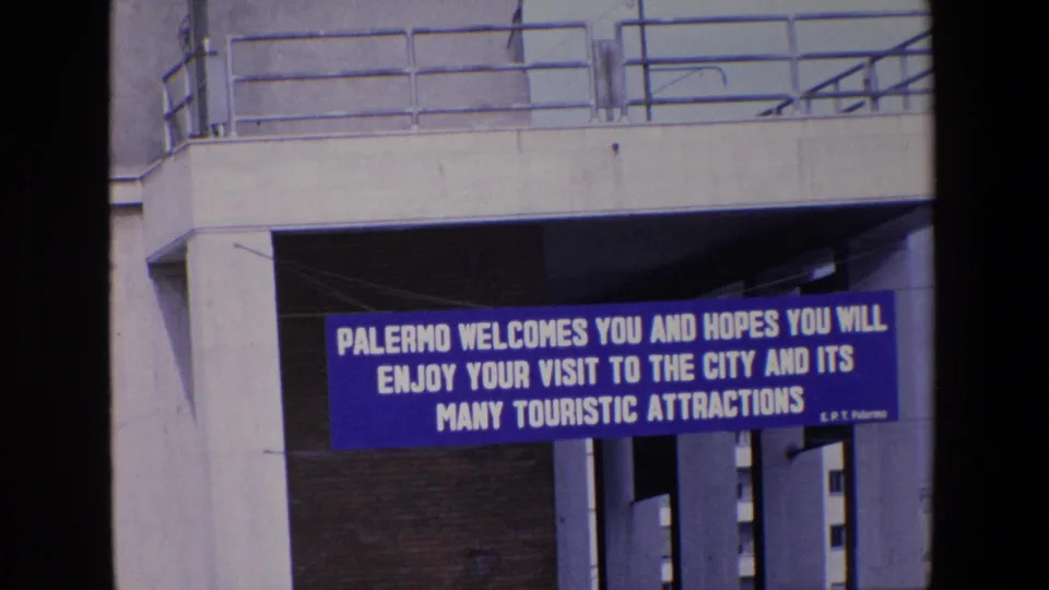 1967 Palermo travel footage frame showing a blue welcome banner with white lettering beneath a concrete overpass and railings.