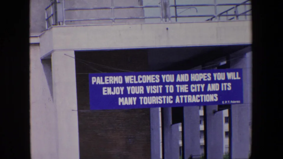 Final 1967 Palermo frame showing the blue tourist welcome banner beneath the concrete overpass, with columns and shadowed spaces behind it.