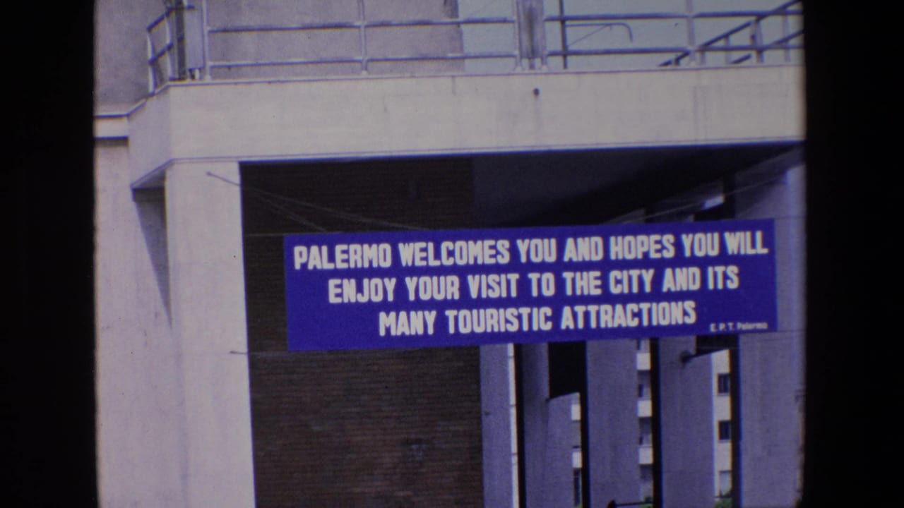 In Palermo, Italy, a blue banner beneath a concrete structure welcomes visitors to the city and its touristic attractions in 1967 home-movie footage.