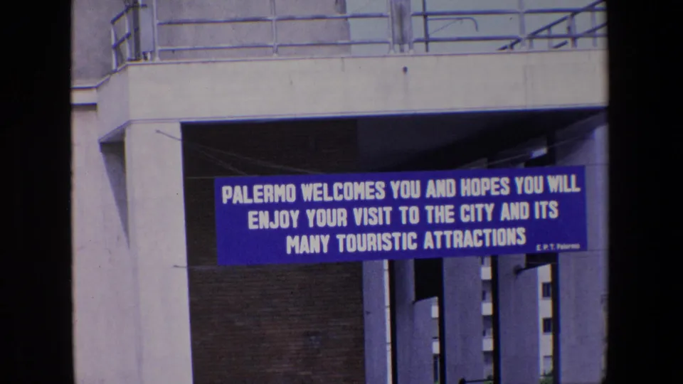In Palermo, Italy, a blue banner beneath a concrete structure welcomes visitors to the city and its touristic attractions in 1967 home-movie footage.