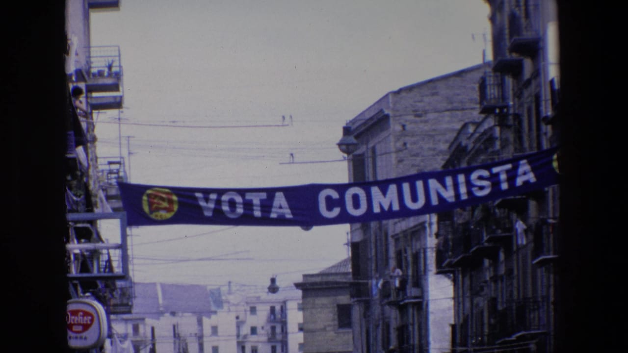 A Vota Comunista political banner with a red and yellow emblem spans a narrow Palermo street lined with balconies, signs, and overhead wires.