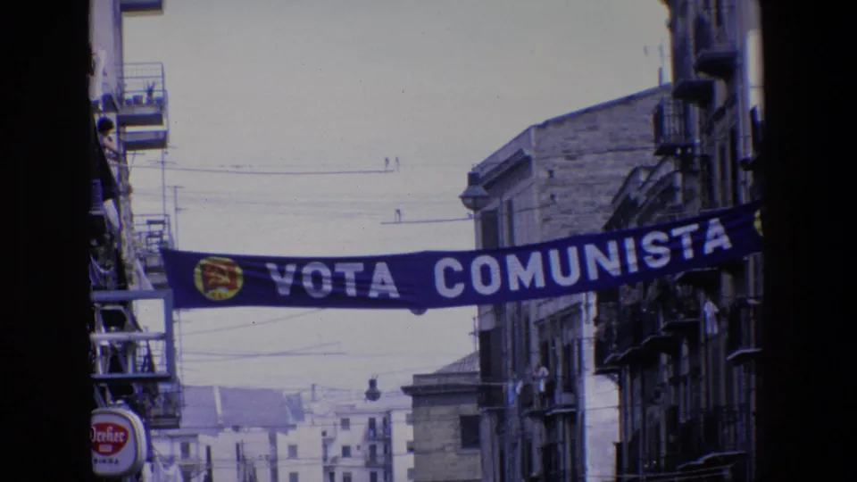 A Vota Comunista political banner with a red and yellow emblem spans a narrow Palermo street lined with balconies, signs, and overhead wires.